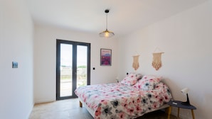 A bedroom with a bed, a desk, a chair, a hanging light fixture, a macrame wall hanging, and a door leading to an outdoor area.