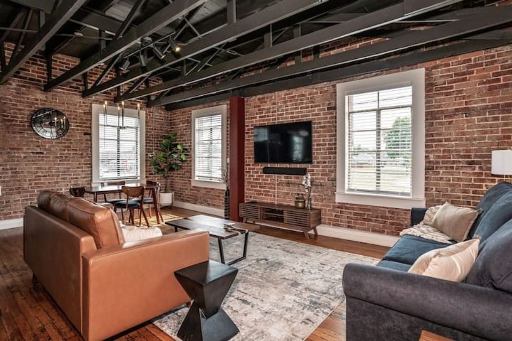 A spacious living room with exposed brick walls, a large flat-screen TV mounted on the wall, and a comfortable seating area with sofas and a coffee table.