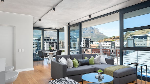 A modern living room with a grey sectional sofa, a round coffee table, and a zebra-patterned ottoman. Large windows offer a view of the city and mountains. - Docklands 502 (Cape Town)