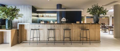 Un área de bar moderna con mostrador de madera, tres taburetes y una planta grande en maceta.