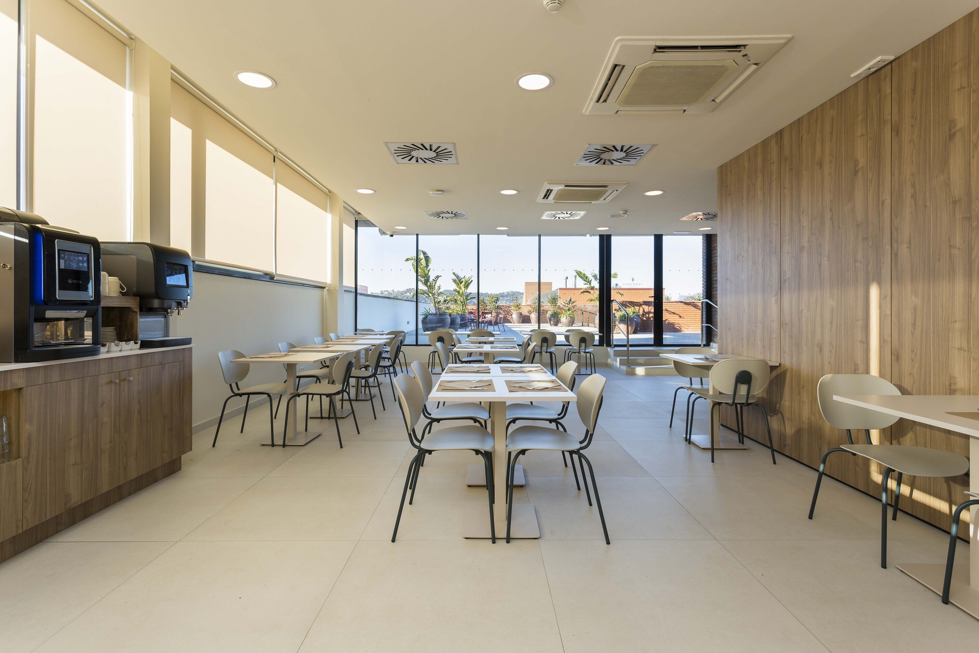 A modern dining area with multiple tables and chairs, a counter with a coffee machine, and large windows offering a view of palm trees and buildings.