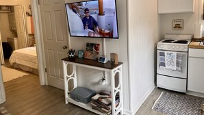 Une pièce confortable avec un téléviseur posé sur un meuble blanc, une petite table avec une valise, et un coin cuisine équipé d’une cuisinière et d’un four.