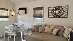 Une pièce dotée d’un plancher en bois blanc, d’un canapé beige agrémenté de coussins à motifs, d’une table à manger blanche avec des chaises, et d’un climatiseur fixé au mur.