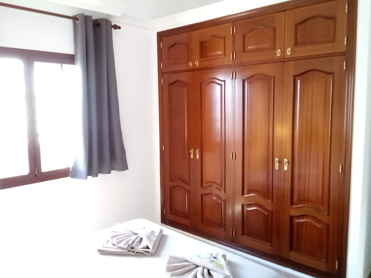 Un dormitorio con un amplio armario de madera, una cama con ropa blanca impecable y una ventana con cortinas grises.