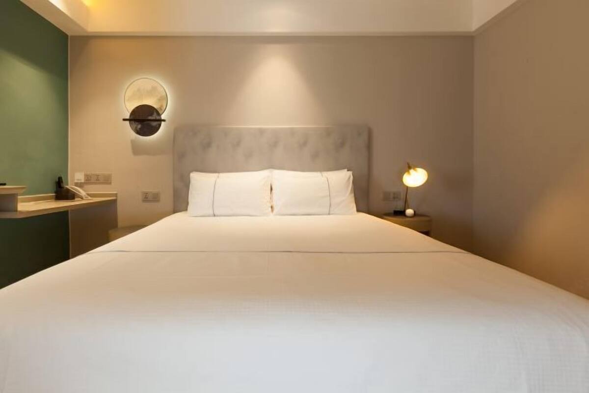 Un lit bien fait, avec une tête de lit, une table de chevet avec une lampe et un luminaire circulaire fixé au mur.