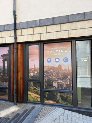 A building with large windows displaying a cityscape and a sign advertising low-cost city apartments with amenities like high-speed WiFi, great communal spaces, and 24/7 guest support. - JOIVY Affordable 3,4,5 and 12 bed private flats next to Royal Mile - Adults only (Edinburgh)