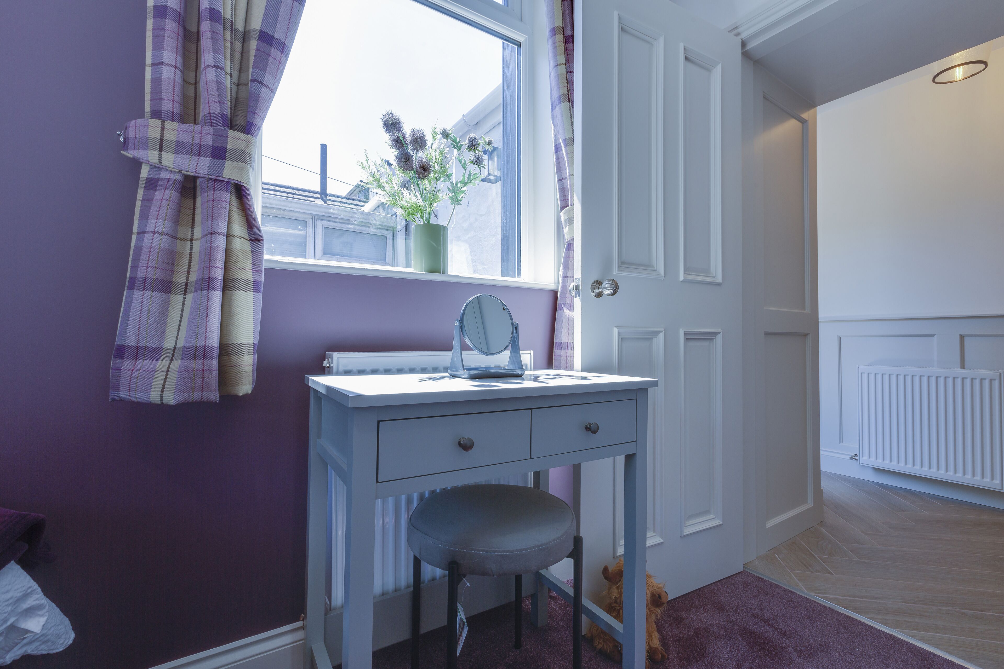 A room with a purple wall, a white desk with a chair, a window with curtains, and a radiator.