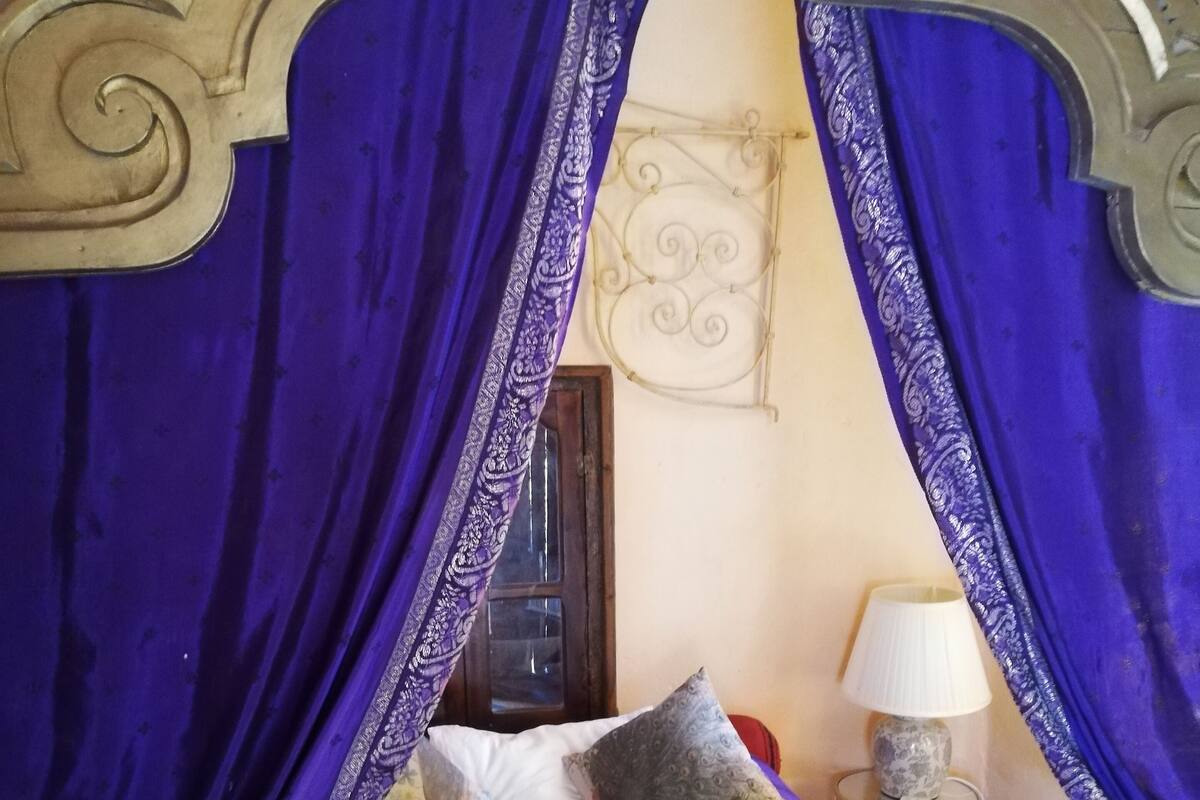 Una cama con una colcha estampada, varios cojines y un dosel con cortinas moradas.