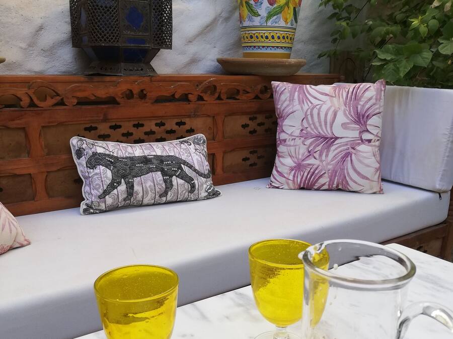 A cozy outdoor seating area with a wooden bench, two cushions, and a table with two yellow glasses and a pitcher of a beverage.