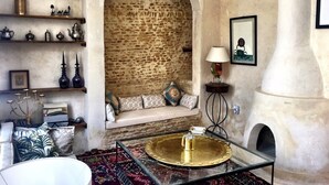 A cozy room with a stone wall, a cushioned bench, a glass table with a tray, and shelves with decorative items. - Riad Dar El Caid. Palais Siecle XIV (Tozeur)