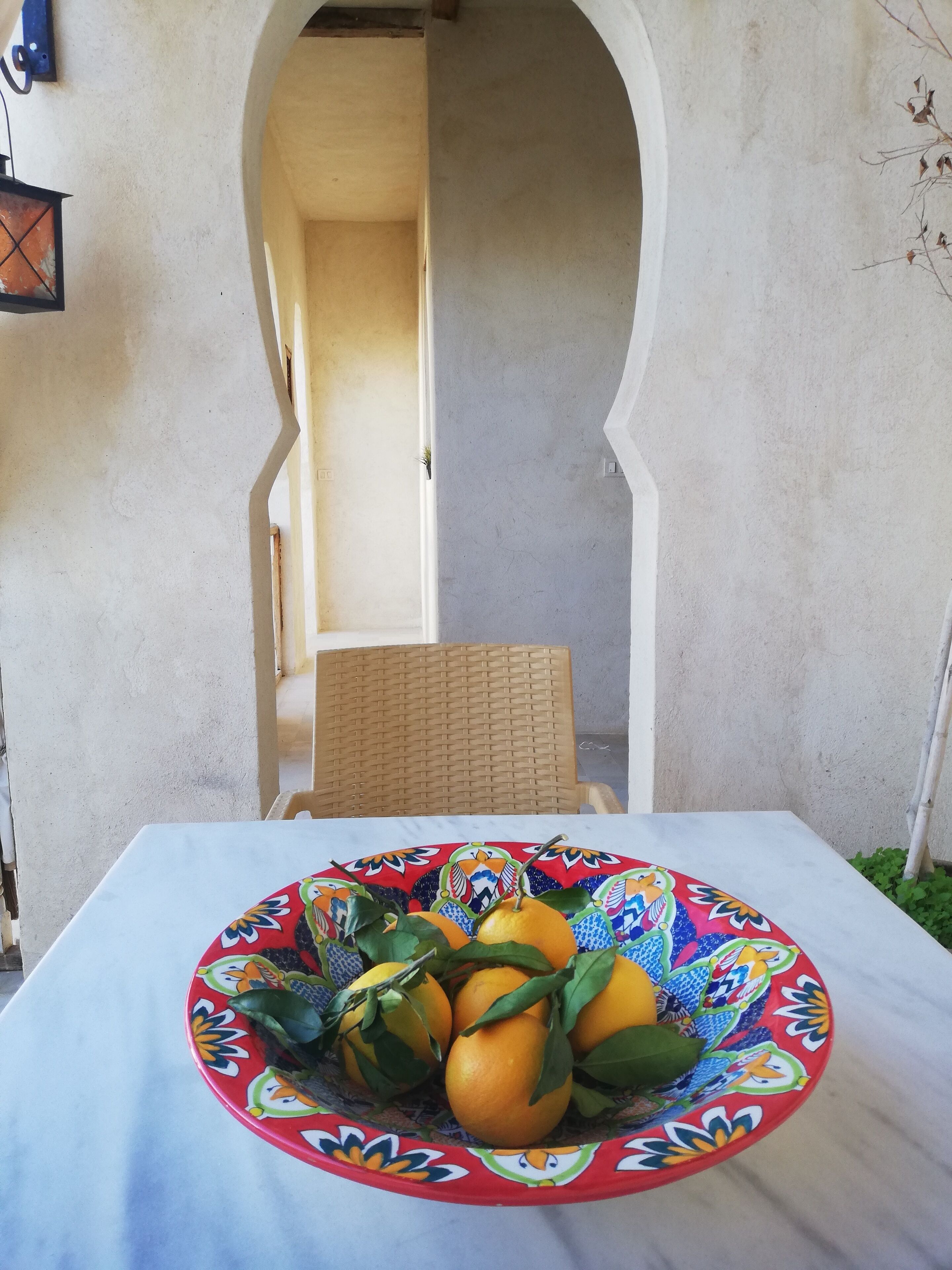 Une table sur laquelle se trouve un panier de fruits colorés, une chaise en osier et un mur blanc avec une porte.