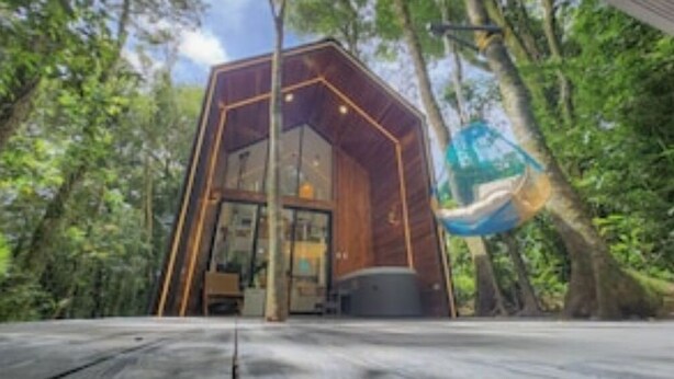 Une cabane moderne en bois, de forme hexagonale, dotée de grandes fenêtres et d’un hamac suspendu à un arbre.