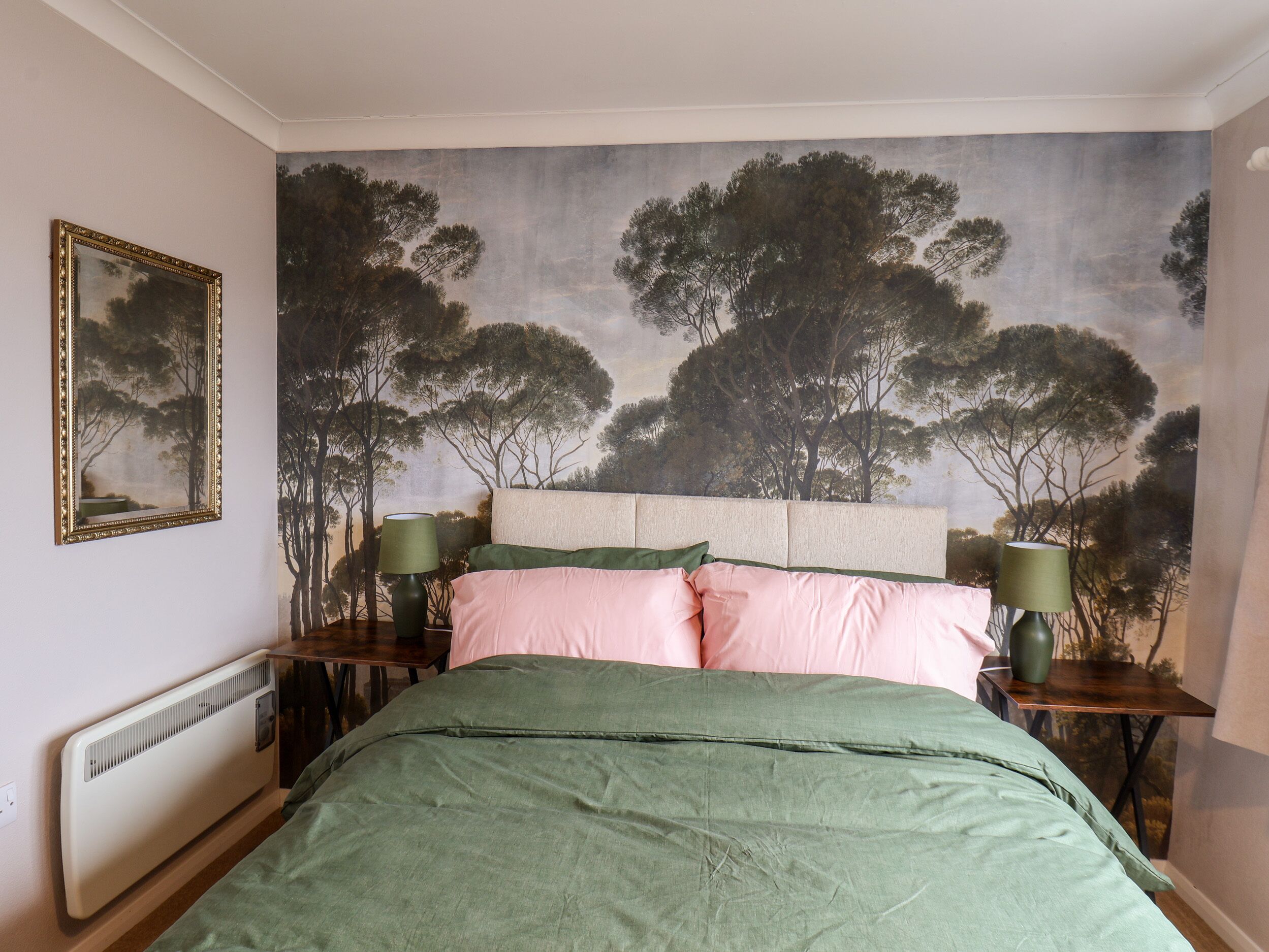 A bedroom with a large bed, a headboard, two bedside tables with lamps, a radiator, and a wall mural of trees.