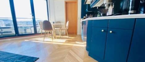 A modern kitchen with blue cabinets, a dining area with a table and chairs, and a large window offering a view of the city.