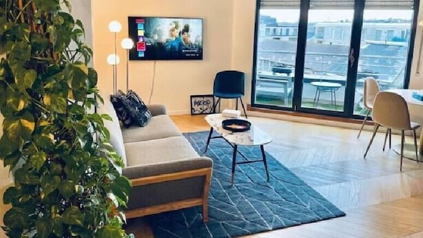 Een moderne woonkamer met een bank, een salontafel en een aan de muur bevestigde televisie. Een groot raam biedt uitzicht op de stadsgezicht.