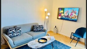 Een woonkamer met een grijze bank, twee gekleurde kussens, een klein rond bijzettafeltje, een blauw vloerkleed, een blauwe fauteuil en een flatscreen televisie aan de muur.