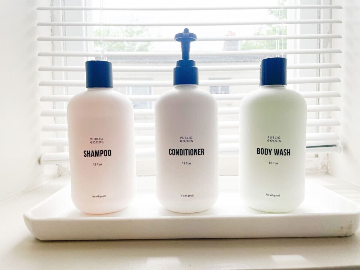 Three bottles of public goods shampoo, conditioner, and body wash on a white tray by a window with blinds.