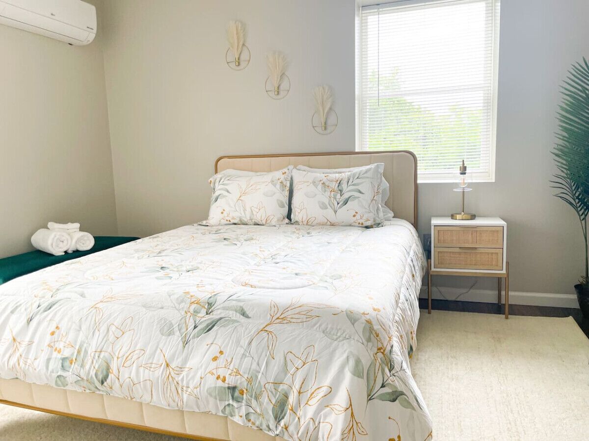 Un lit bien fait, recouvert d’une couvre-lit à motifs floraux, une table de chevet avec une lampe et une plante en pot dans une chambre avec une fenêtre.