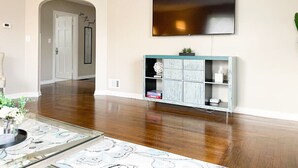 Un vaste salon doté d’un plancher en bois, d’un grand téléviseur à écran plat fixé au mur, d’un meuble bleu clair avec des étagères et d’un tapis décoratif.