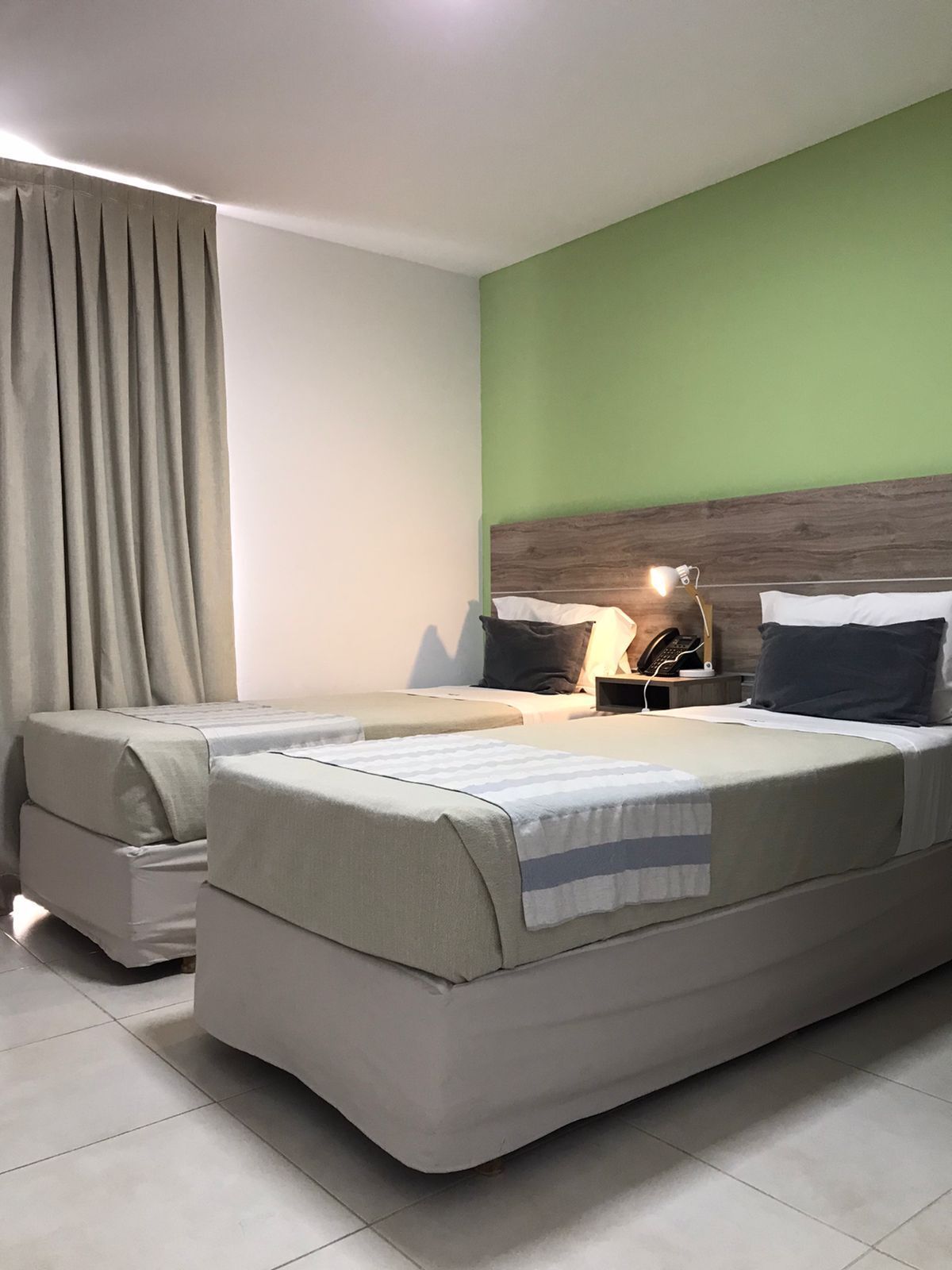 Una cama doble con cabecera de madera, dos mesitas de noche con lámparas, una mesita auxiliar con un teléfono y una ventana con cortinas.