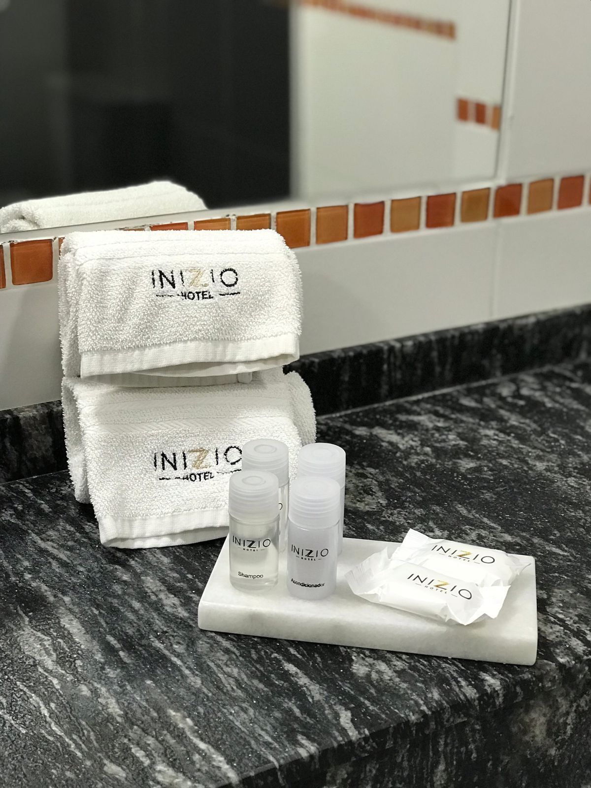 A bathroom with a marble countertop, two stacked white towels, and three toiletries labeled 'INIZIO HOTEL'.