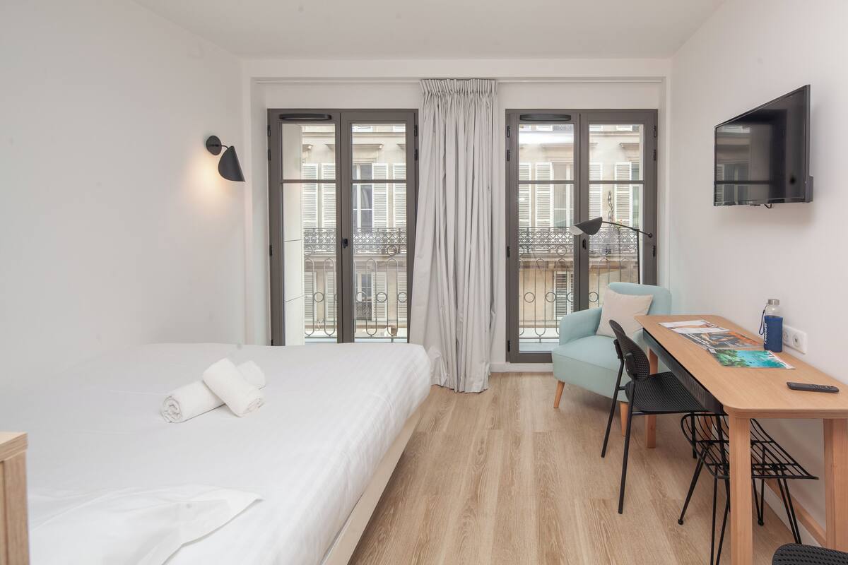 Habitación de hotel con cama, escritorio, silla, televisión y balcón con vistas a edificios.
