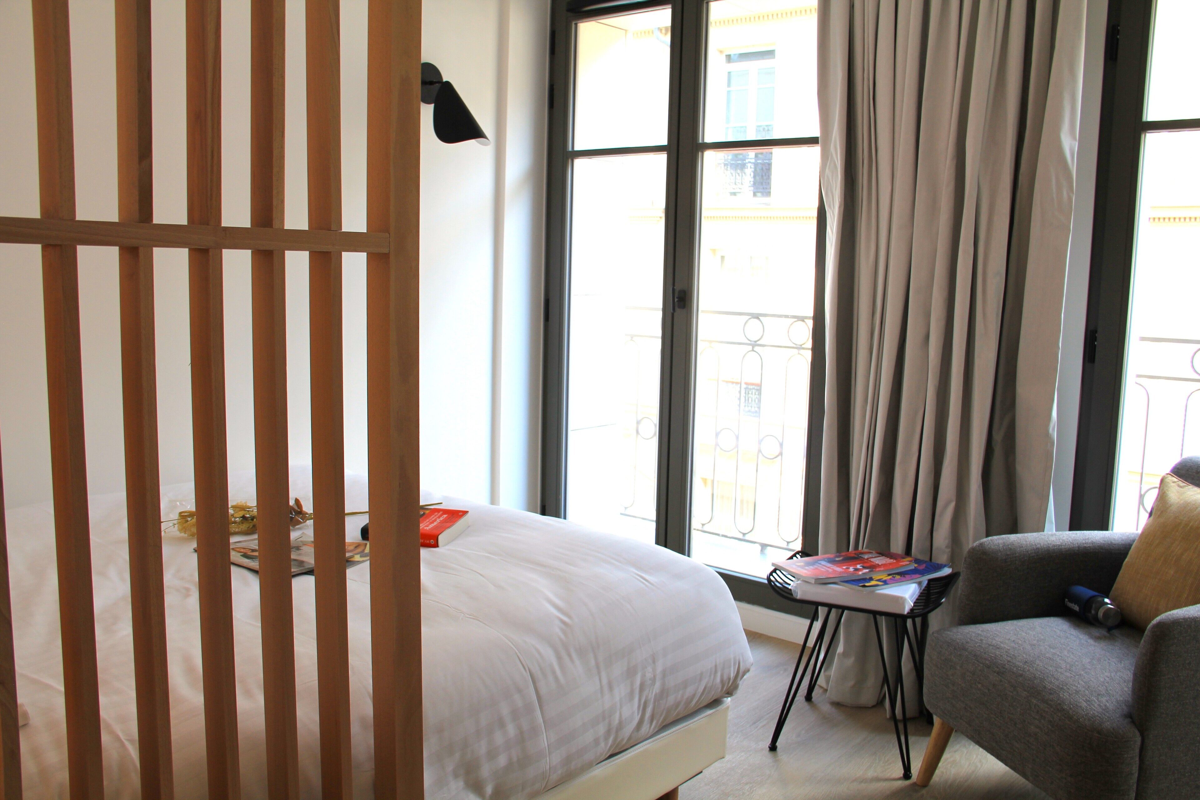A bedroom with a bed, a chair, a small table with books, and a window with curtains.