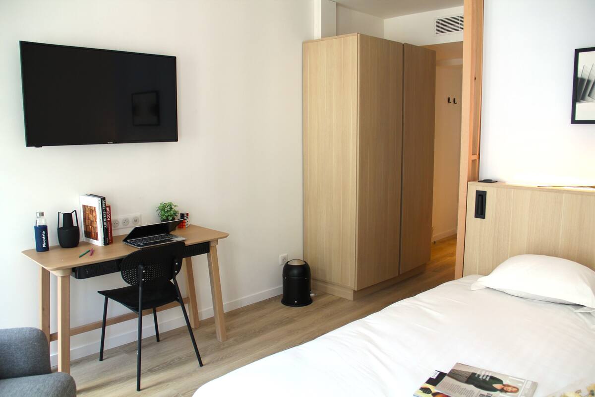 Habitación de hotel con una cama, un escritorio con una computadora portátil, una silla, un armario y un televisor en la pared.