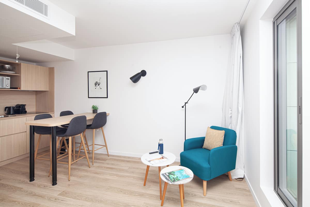 Una sala de estar moderna con un sofá azul, una mesa de comedor con sillas, una pequeña mesita auxiliar y una lámpara de pie.