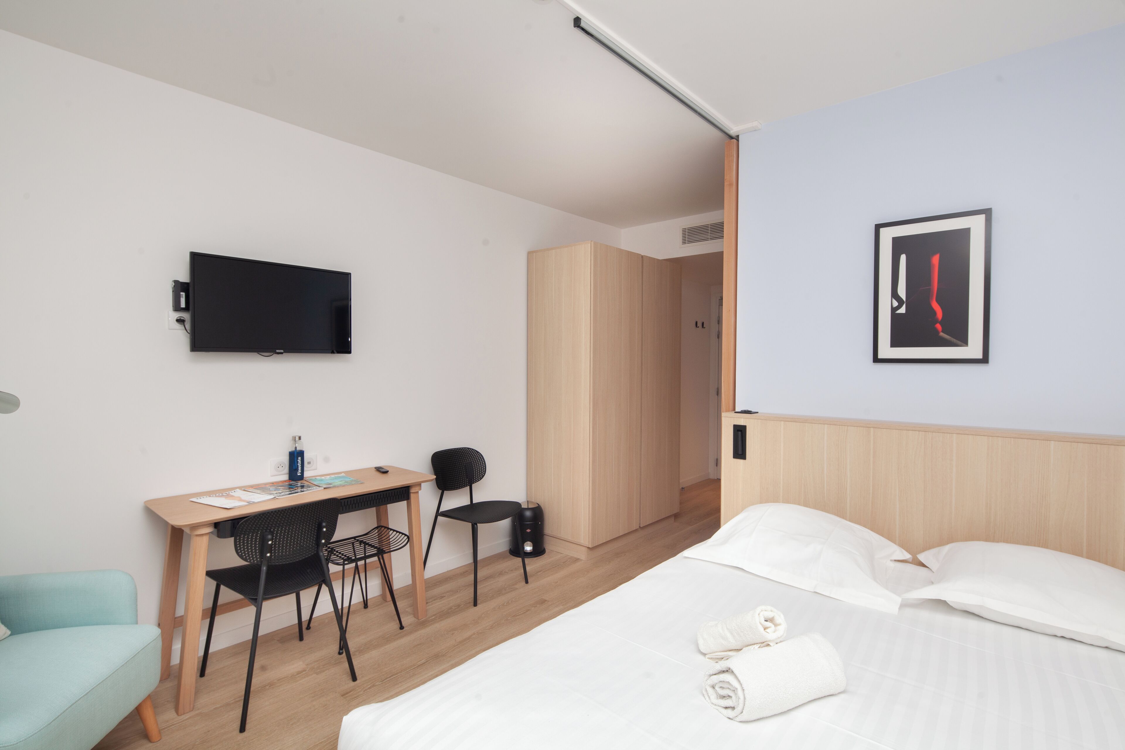 A modern hotel room with a bed, a desk with a chair, a television mounted on the wall, and a framed picture on the wall.