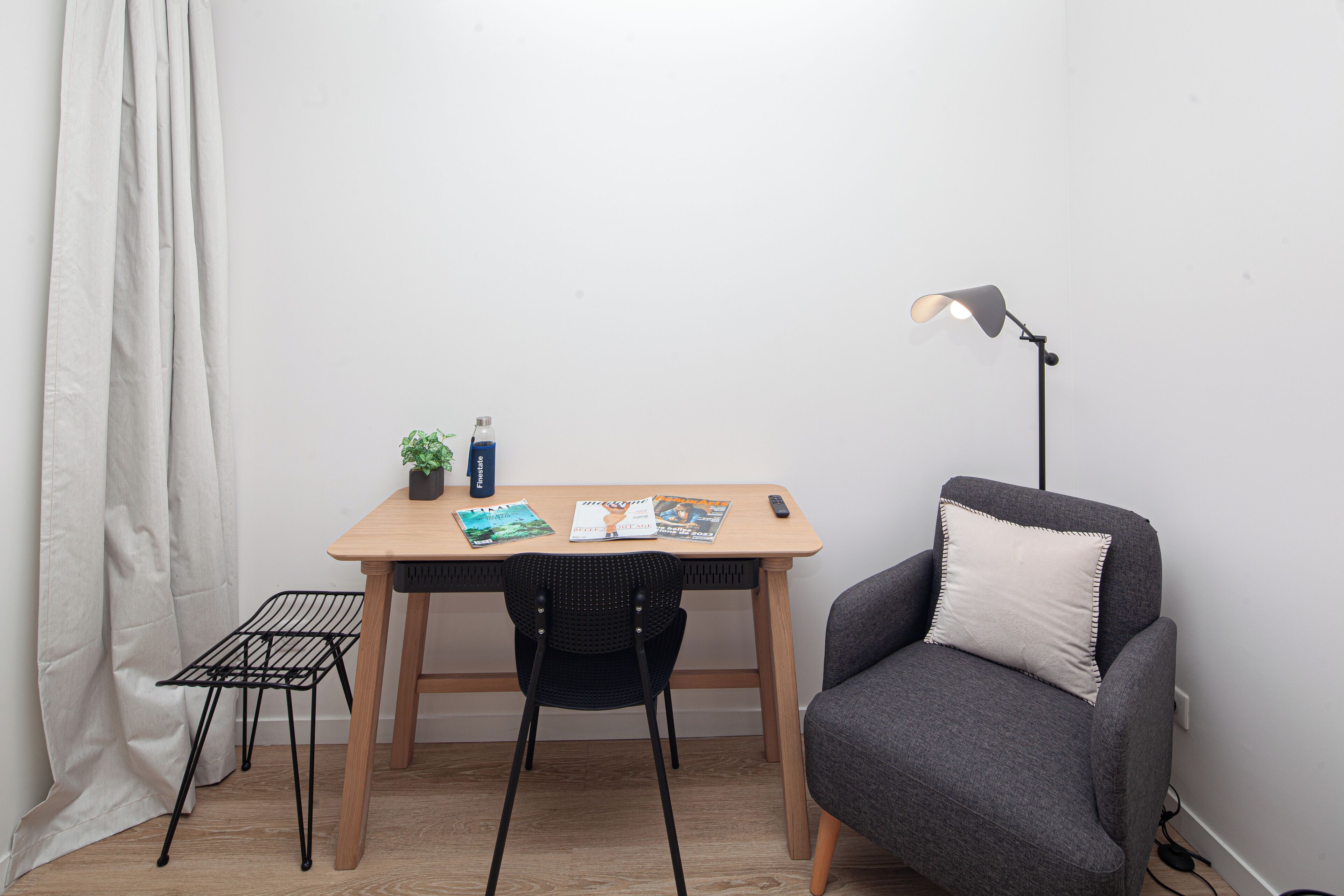 Ein kleines Zimmer mit einem Holztisch, einem schwarzen Stuhl, einem grauen Sofa, einer Stehlampe und einem Vorhang.