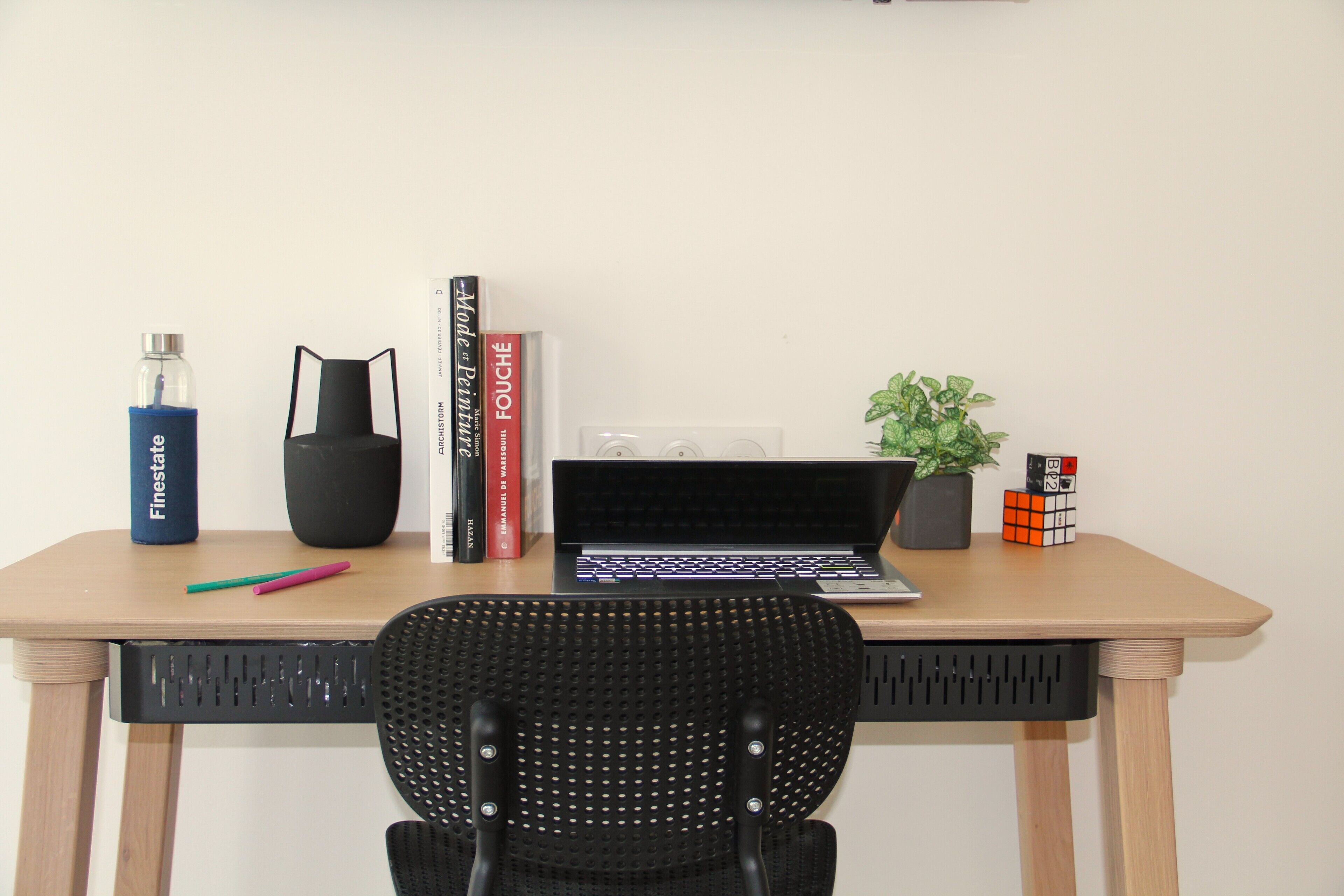A wooden desk with a laptop, a black chair, a potted plant, a Rubik's cube, a water bottle, and a few books.