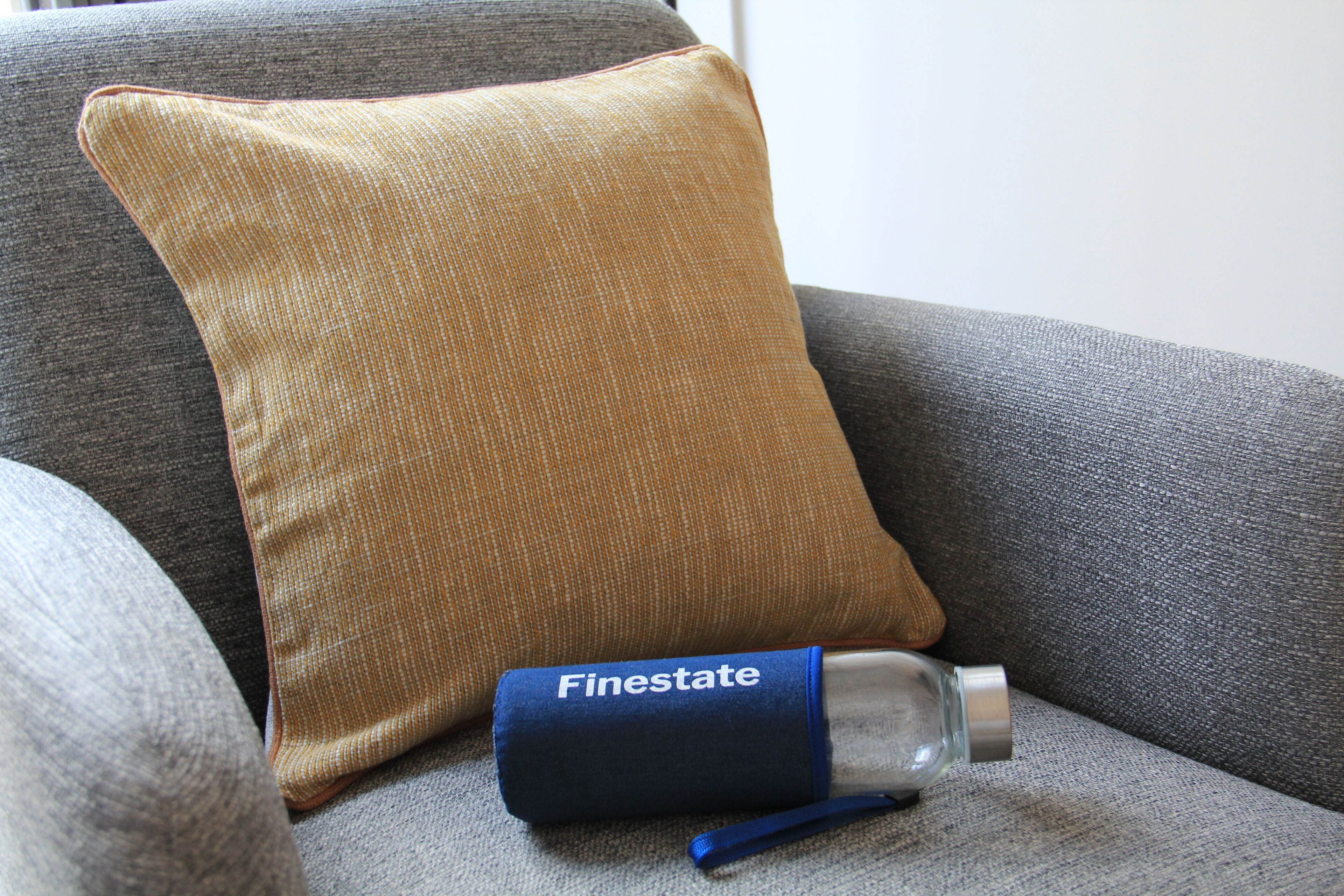 A grey sofa with a mustard-colored pillow and a bottle labeled "Finestate" on it.