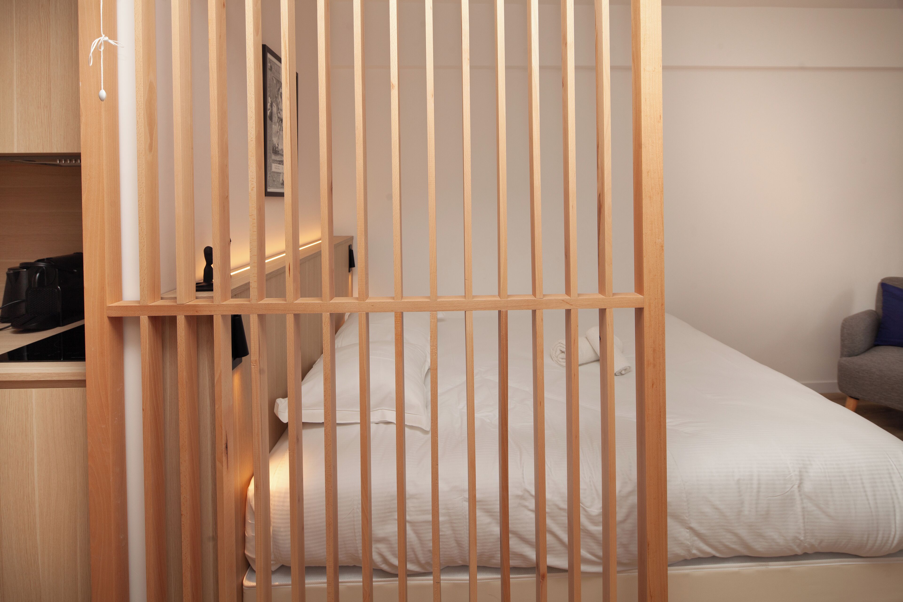 A bedroom with a wooden partition, a bed with white bedding, a grey armchair, and a small shelf with items.