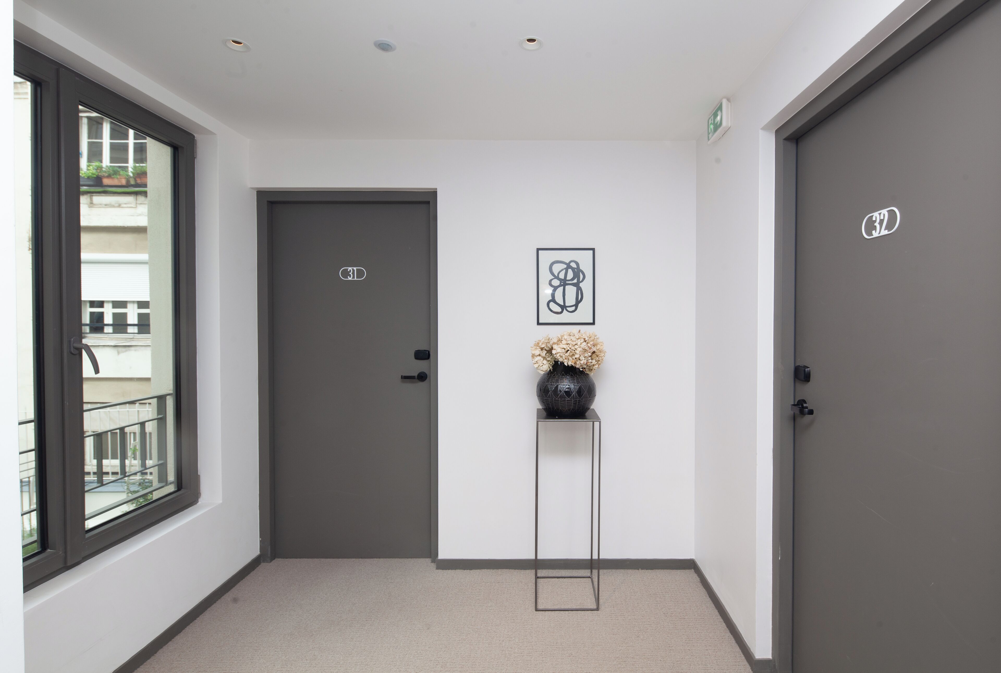A hallway with a grey door, a framed artwork, and a vase with flowers on a pedestal.
