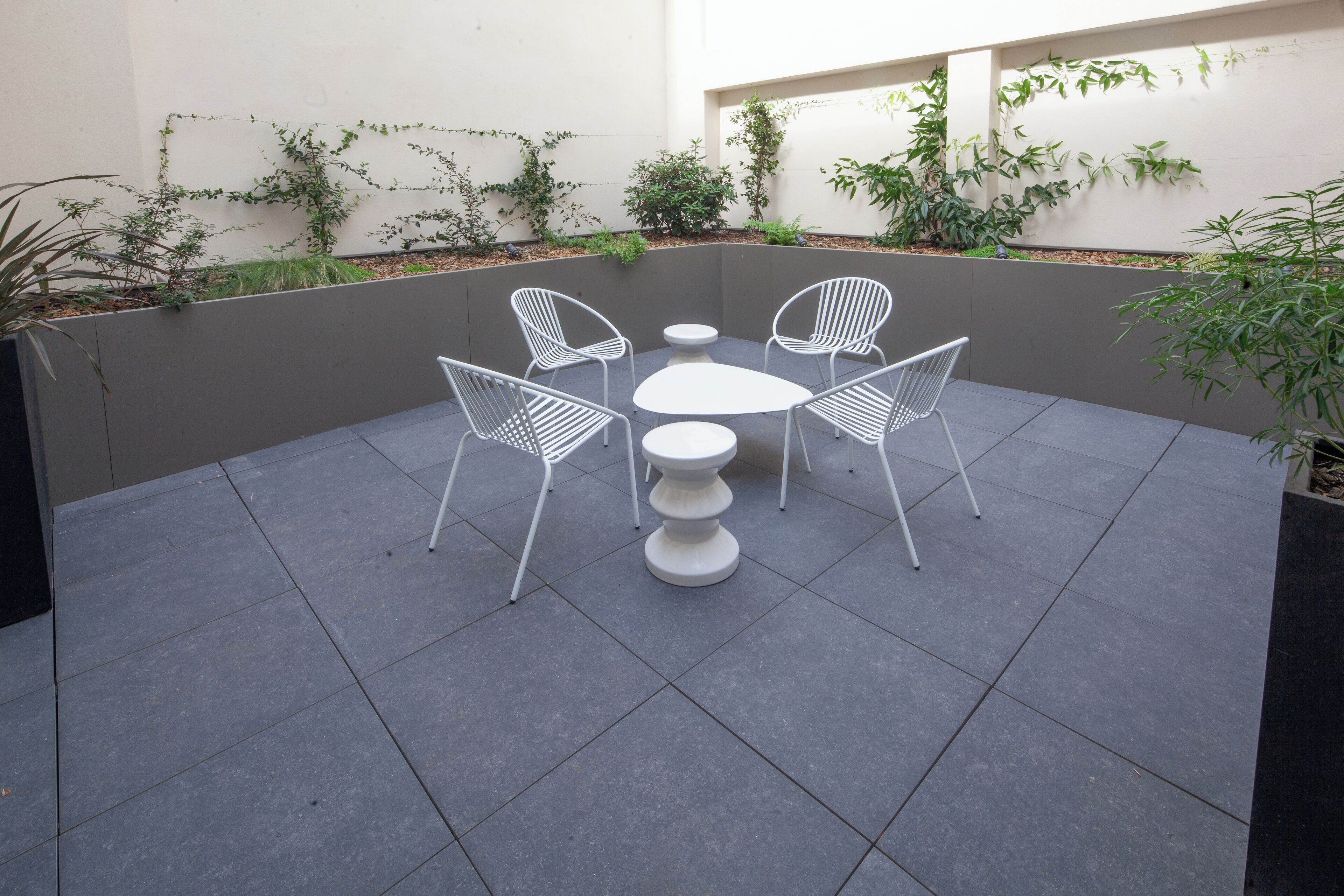 Un patio con una mesa redonda y cuatro sillas, rodeado de plantas y un muro bajo.
