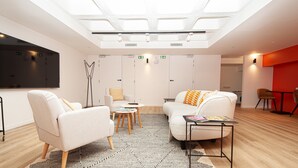 Une pièce moderne et bien éclairée, avec un plafond haut, des murs blancs et un grand téléviseur à écran plat. On y trouve deux canapés blancs avec des coussins orange, un lampadaire et une petite table ronde sur laquelle sont posés des magazines.