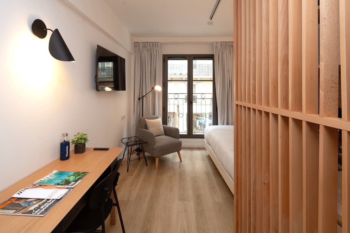 Un dormitorio pequeño con una cama de madera, un escritorio con silla, una mesita con una planta y un televisor montado en la pared.