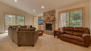 A spacious living room with a stone fireplace, two sofas, and large windows offering a view of trees.