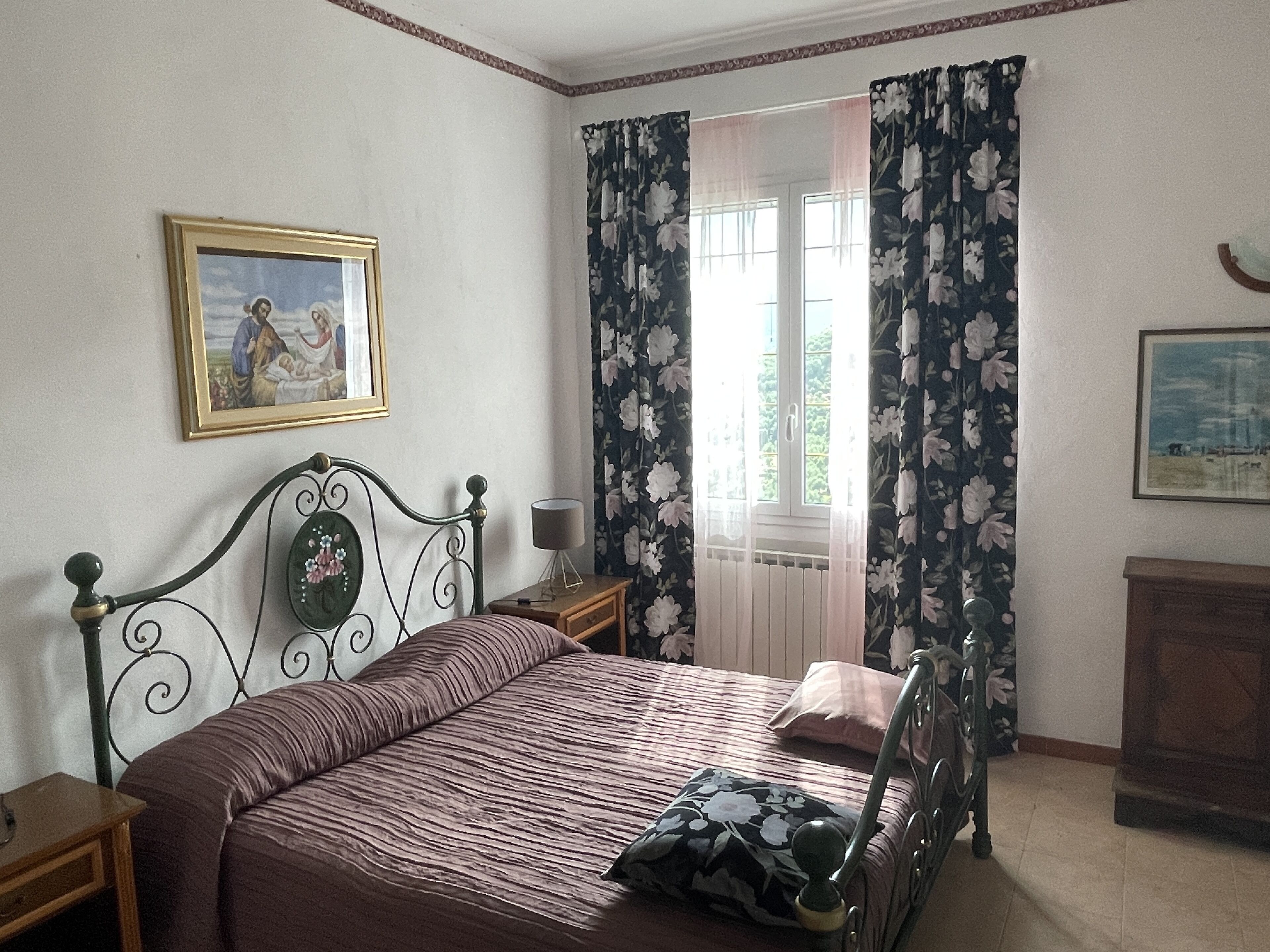 A bedroom with a bed, a nightstand, a lamp, a window with floral curtains, and two framed pictures on the wall.