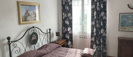 A bedroom with a bed, a nightstand, a lamp, a window with floral curtains, and two framed pictures on the wall.