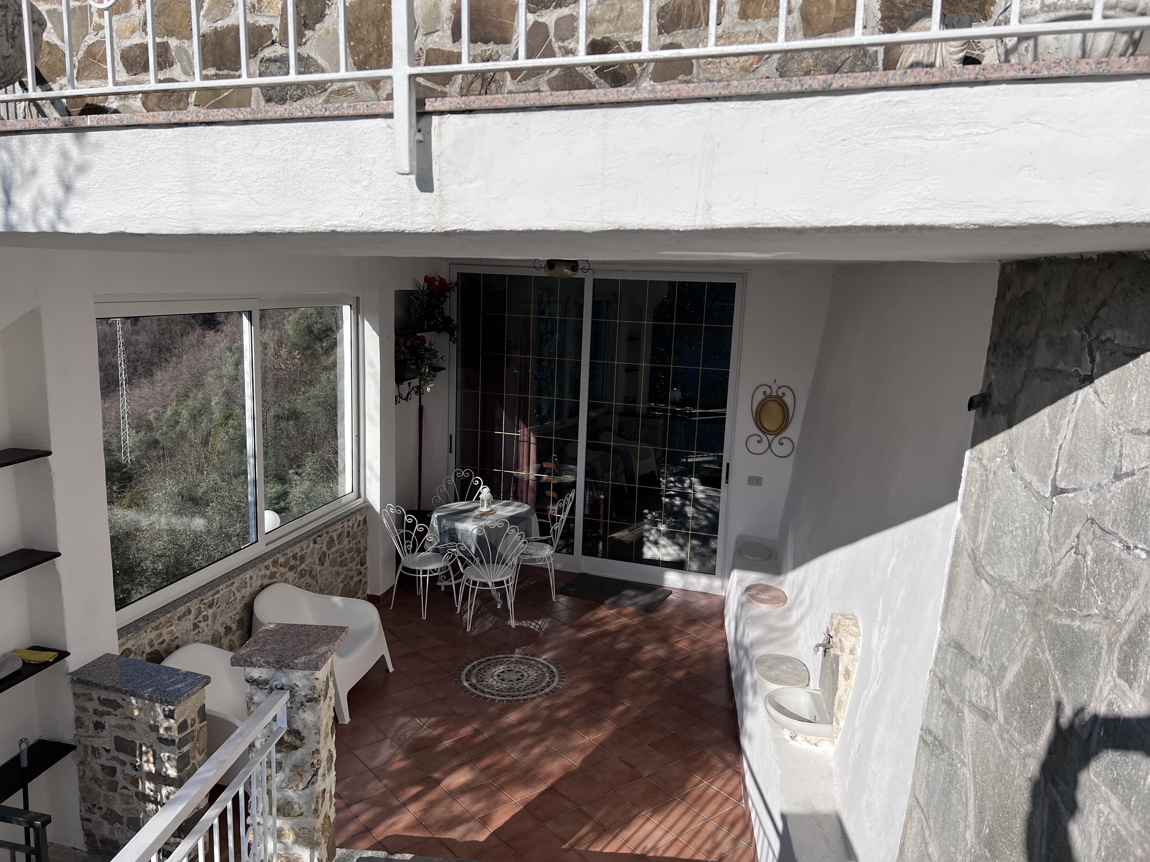 Une terrasse ensoleillée avec des murs en pierre, un coin repas avec des chaises blanches et une vue à travers une porte vitrée.