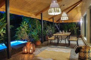 Un área de comedor al aire libre con piscina, plantas en macetas e iluminación colgante.