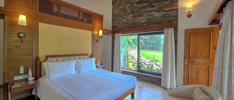 A bedroom with a large bed, wooden headboard, a ceiling fan, a stone wall, a window with a view of a garden, and a door.