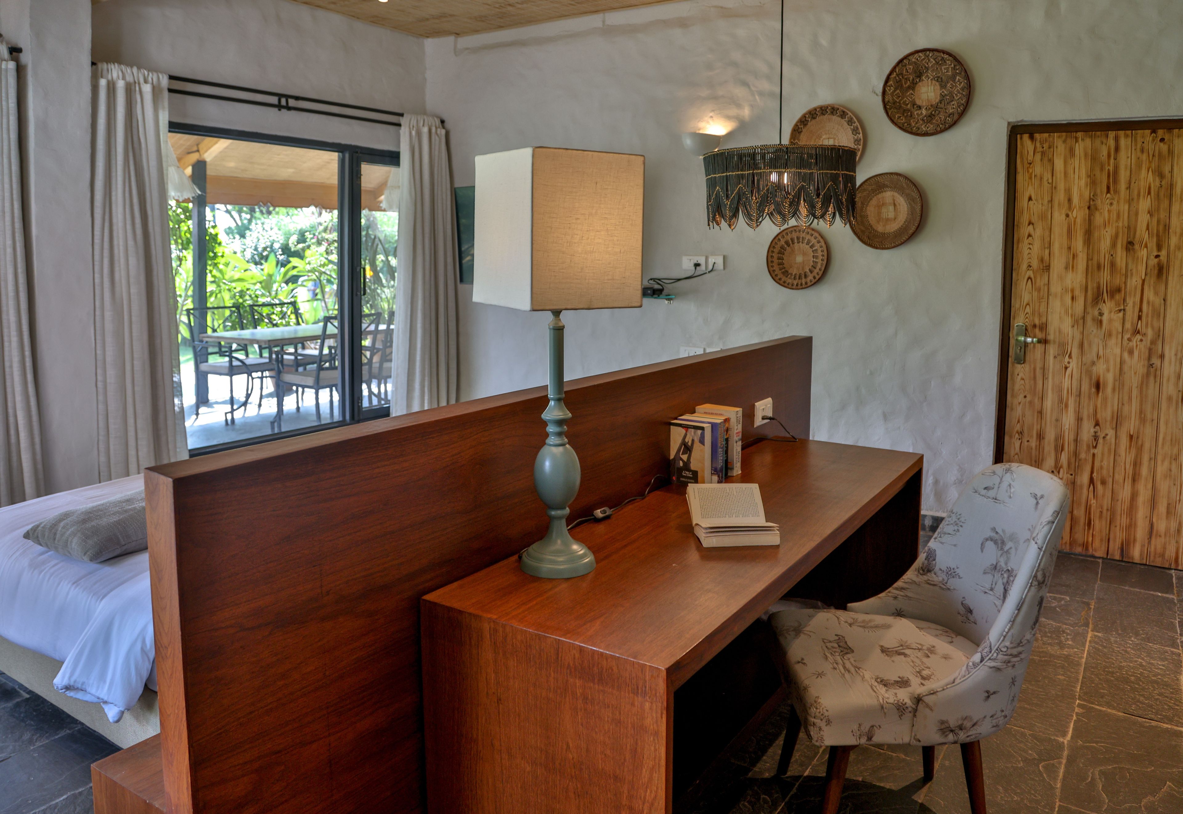 A hotel room with a wooden desk, a lamp, a chair, a bed, and a view of the outdoors.