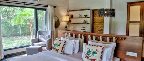 Habitación de hotel con una cama grande, una silla, una mesita de noche, una lámpara, una estantería con adornos y vistas al exterior.