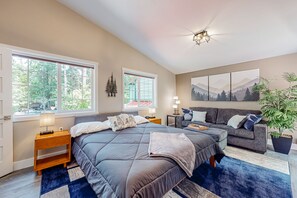 A bedroom with a large bed, a sofa, two bedside tables, a window with a view of trees, and a wall-mounted artwork. - Rare, dog-friendly riverfront retreat with hot tub access plus kayaks & AC (Tahuya)