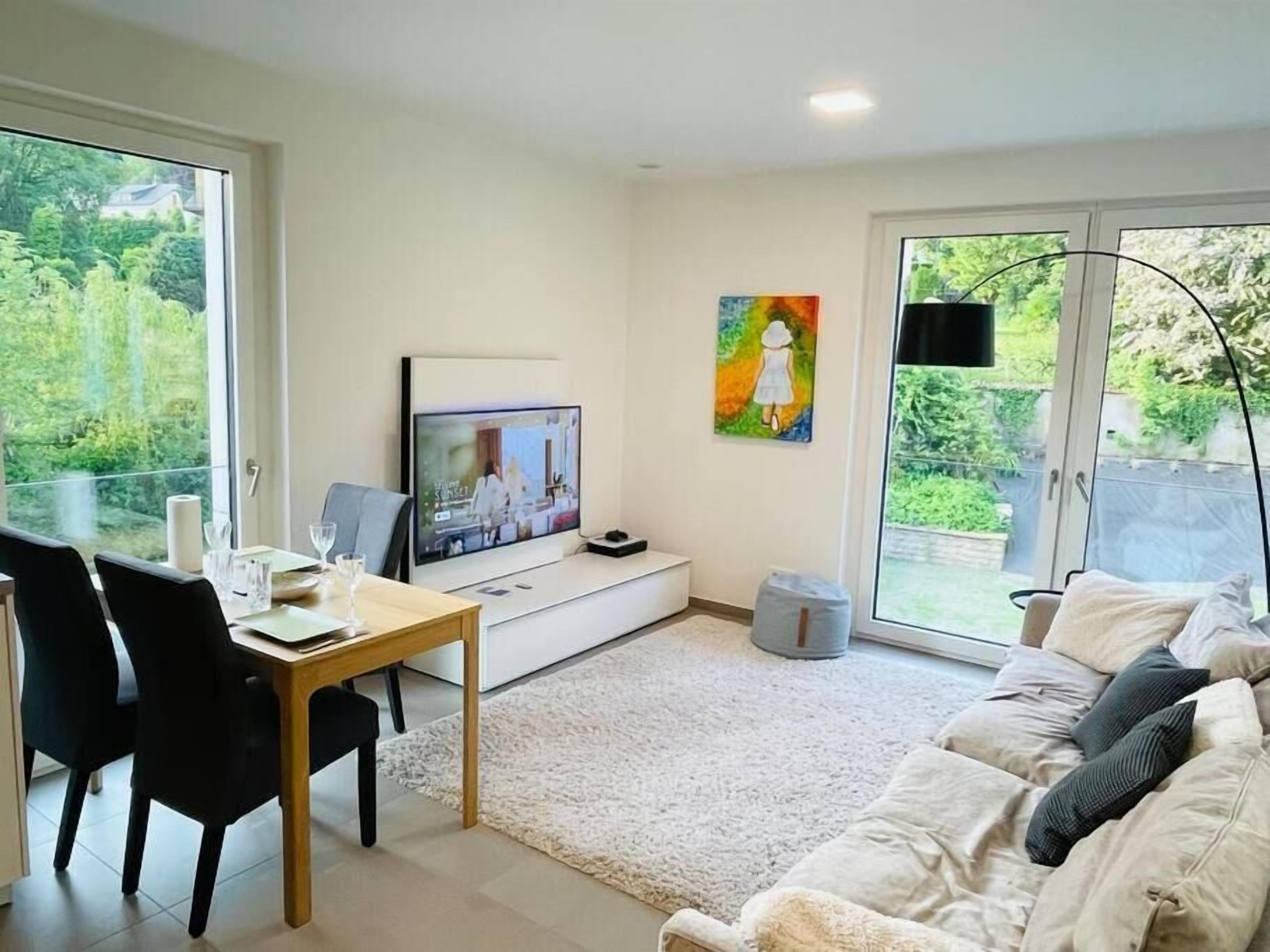 A modern living room with a TV, dining area, and large windows overlooking greenery.