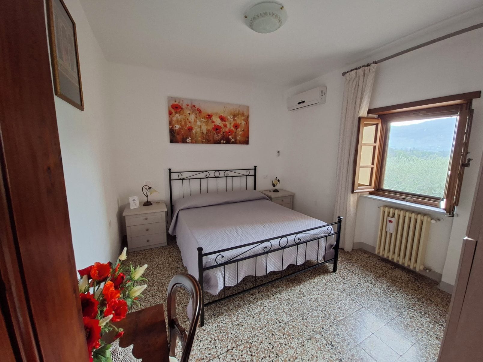 Un dormitorio con cama, mesita de noche, radiador, ventana con vistas y un cuadro en la pared.
