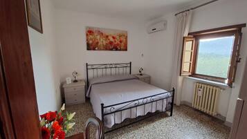 Un dormitorio con cama, mesita de noche, radiador, ventana con vistas y un cuadro en la pared.